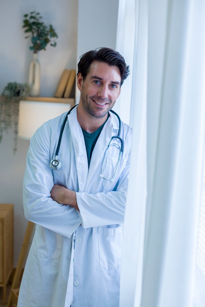 Smiling male doctor wearing a white coat and stethoscope stands by a window inside a home medical setting, Technology, young adult.