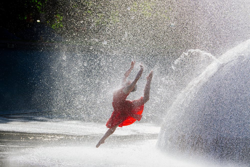 Female dancer in a red dress leaps mid-air through the spray of a large outdoor fountain under sunlight, USA.