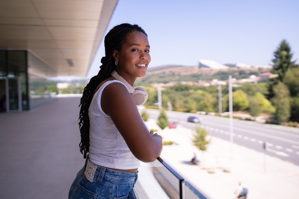 A young woman with headphones around her neck smiles while standing on an outdoor balcony overlooking a road and greenery, remote work, travel, Santiago De Compostela, Spain.