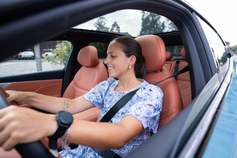 Smiling woman with black hair and a tattoo drives a modern car with leather seats and an open sunroof during daytime, Spain.