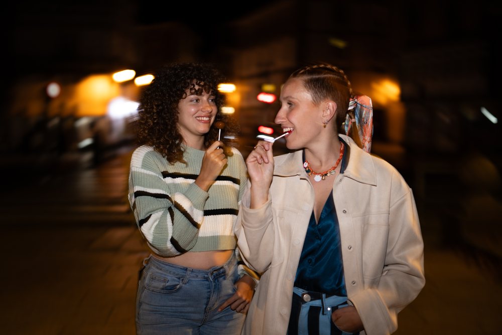 Two women smiling and holding lollipops on a nighttime urban street, friendship, Santiago De Compostela, Spain.