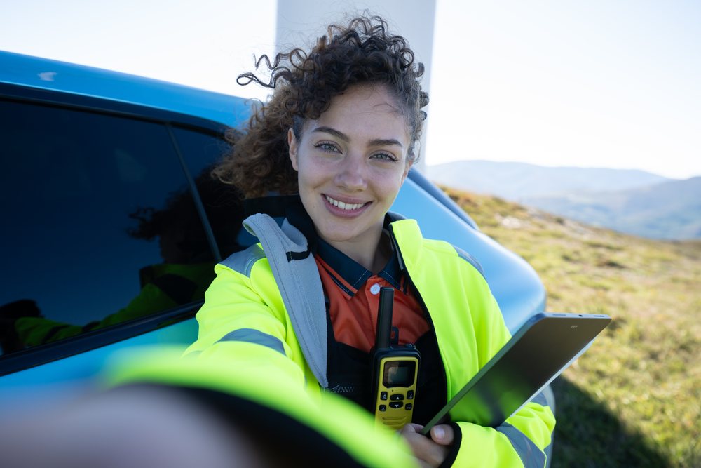 Woman wearing a high-visibility jacket holding a tablet and walkie-talkie outdoors with mountainous scenery, sustainability, renewable energy, Galicia, Spain.