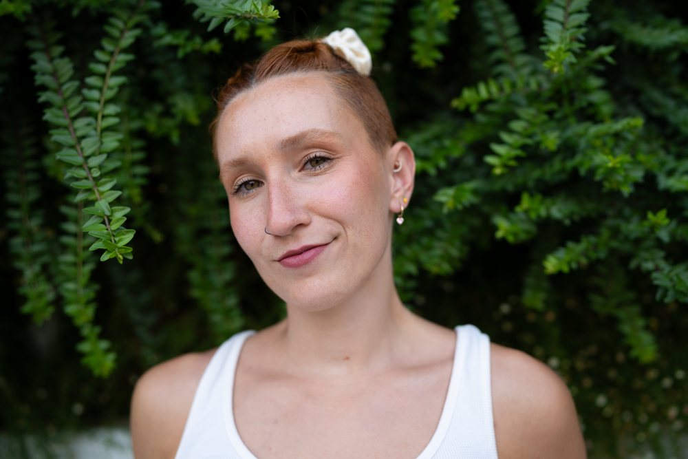 A confident young woman with short hair and a scrunchie smiles outdoors against a backdrop of green plants, urban, Santiago De Compostela, Spain.