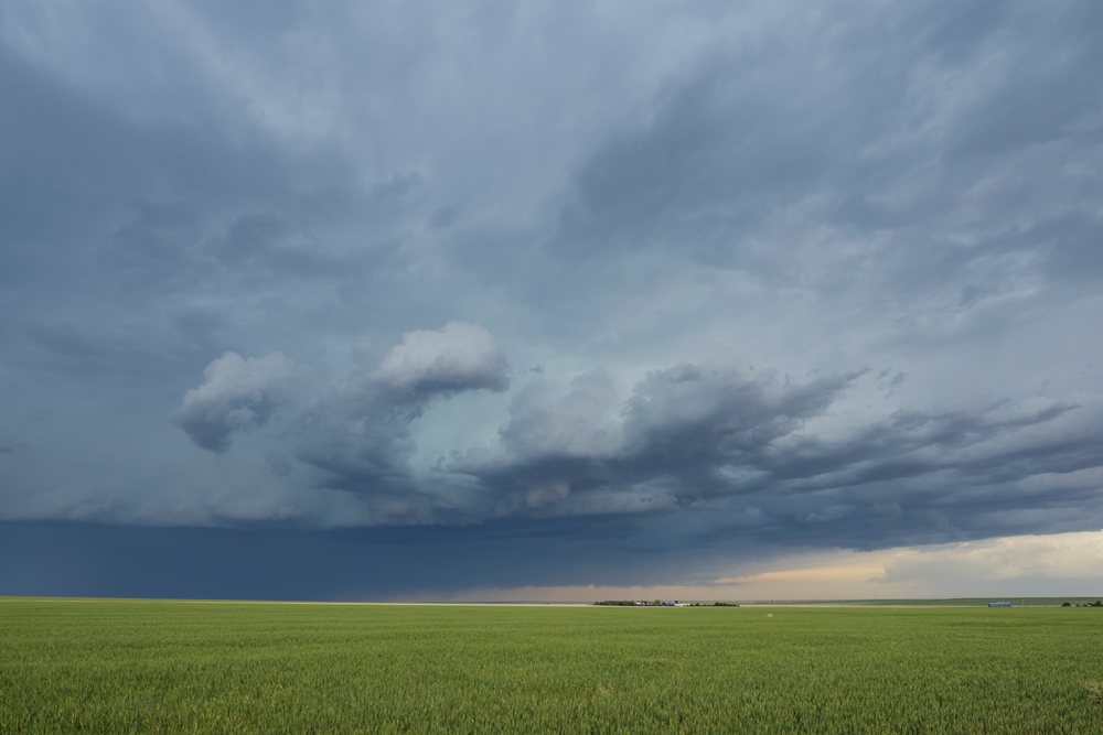 Storm clouds gather above a flat green grassland with farm buildings visible on the horizon on the Great Plains, Storm Chasing, Weather, Environmental, Lindon, Colorado, USA.