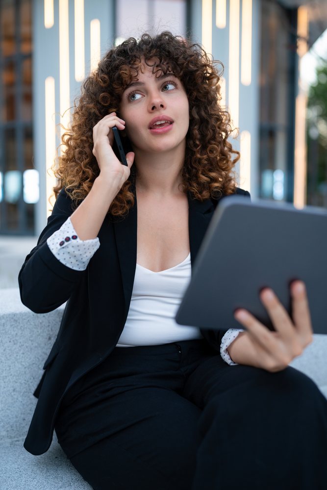 Young woman in a black blazer talking on a phone while holding a tablet outdoors, Madrid.