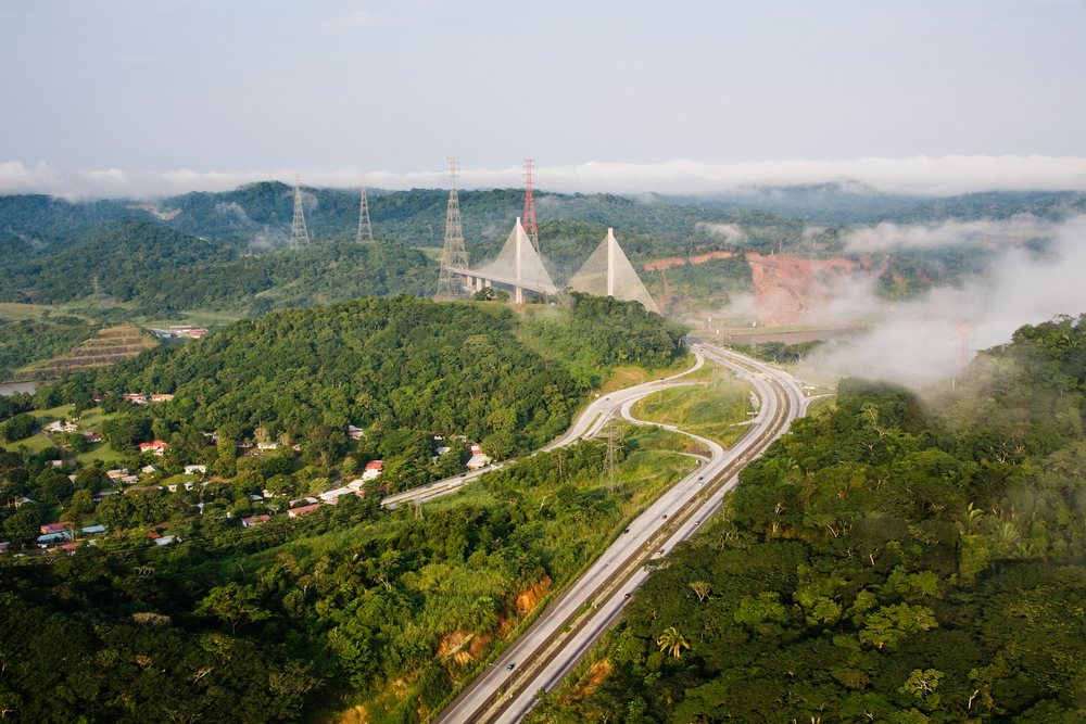 Panama Canal Bridge spans over a highway amidst tropical forest and misty mountainous terrain with transmission towers nearby.