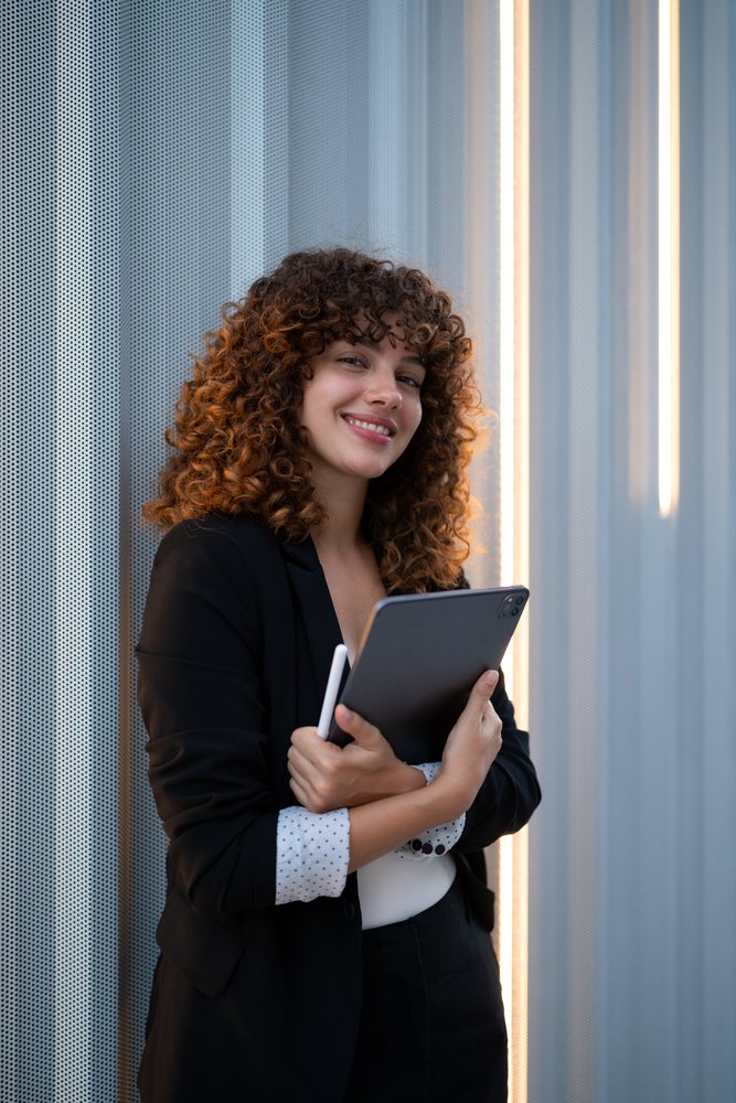 Young woman with curly hair, holding a tablet and digital pen, smiling in a modern office with a gray perforated wall and vertical light strip, Madrid.