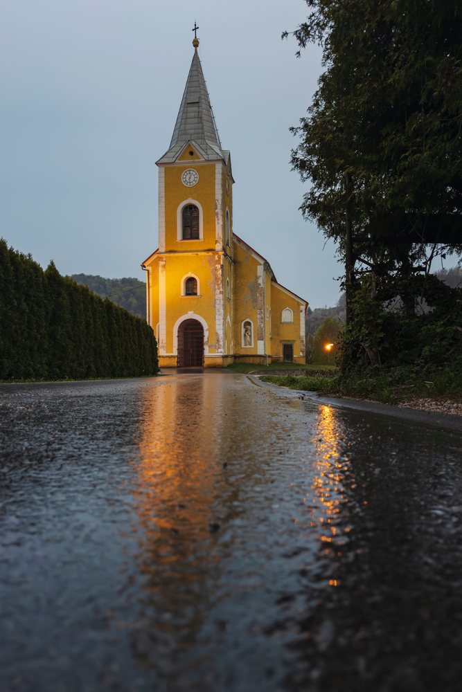 Yellow-facade Church of St Valentine with clock tower and cross reflects on wet road, framed by hedges and trees at evening, landscape, cityscape, Žusem, Slovenia.