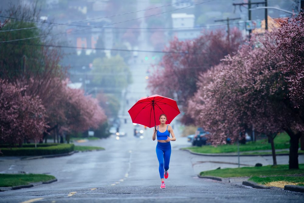 Woman running outdoors holding a red umbrella on a rainy urban street lined with blossoming cherry trees, USA.