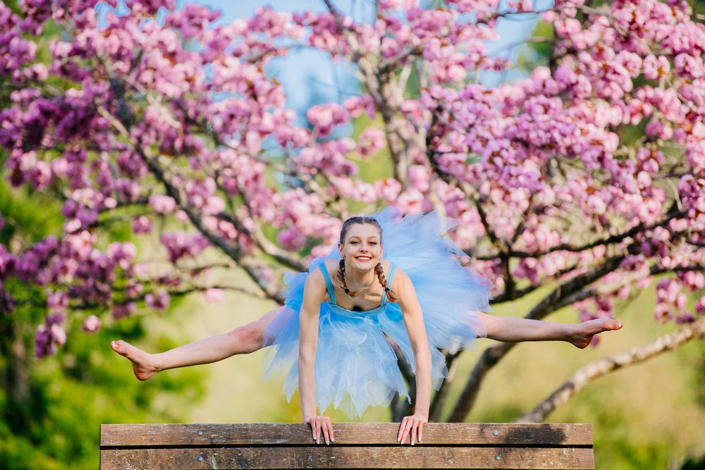 Ballet dancer in a blue tutu performs a split jump over a bench in a park amidst pink blossoming trees in spring, USA.