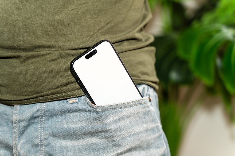 Smartphone with white screen placed in the front pocket of blue jeans worn with a green shirt indoors, mock up, Spain.