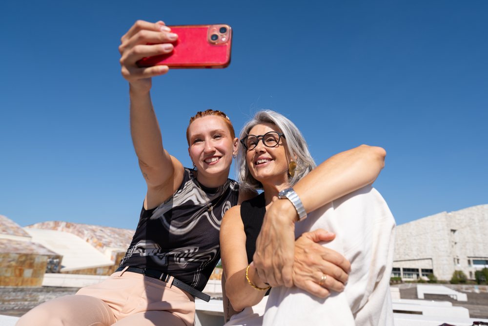 Two women smiling and taking a selfie outdoors in bright sunny weather with modern urban architecture, businesswomen, Spain.