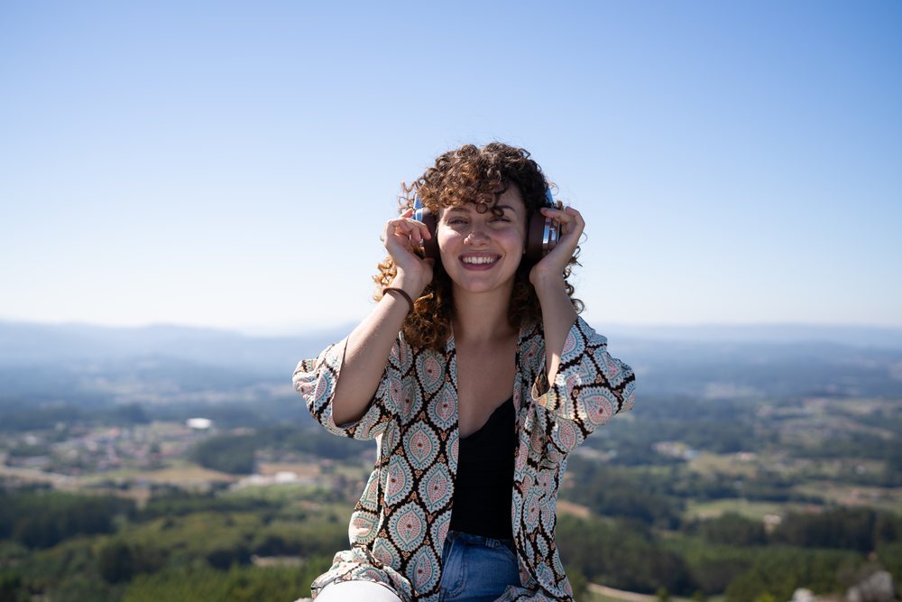 Smiling woman with curly hair listens to music on headphones outdoors at an elevated viewpoint overlooking, Pico Sacro, Galicia, Spain.