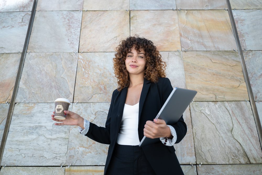 Woman holding a coffee cup and laptop standing against a stone wall outdoors, businesswoman, traveling executive, Spain.