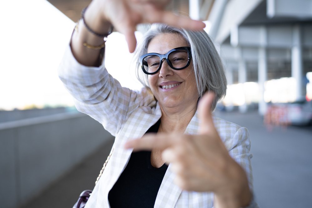 Mature woman with gray hair wearing glasses and a checkered blazer smiles while framing the scene with her hands outdoors in an urban setting, businesswoman, Spain, Madrid.