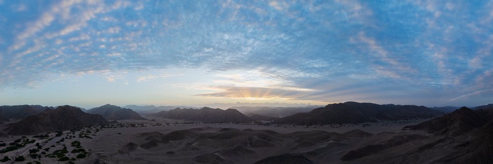 Sunrise illuminates the mountainous Hoanib River Valley in Namibia's Kunene Region, revealing a serene desert landscape with clouds and sparse vegetation.
