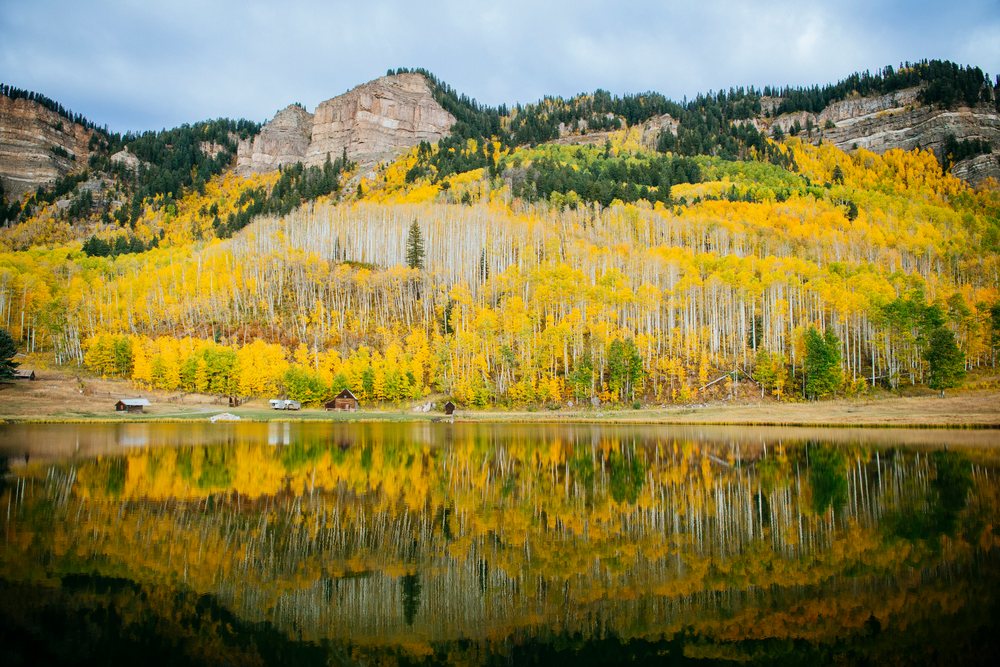 Yellow aspen trees in autumn colors reflect on a calm mountain lake with wooden cabins nestled on the hillside, Durango, Colorado, USA.