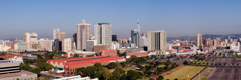 Skyline of Durban city with clustered high-rise buildings, visible greenery, urban park, and a large parking lot under a clear blue sky, Kwazulu Natal, South Africa.