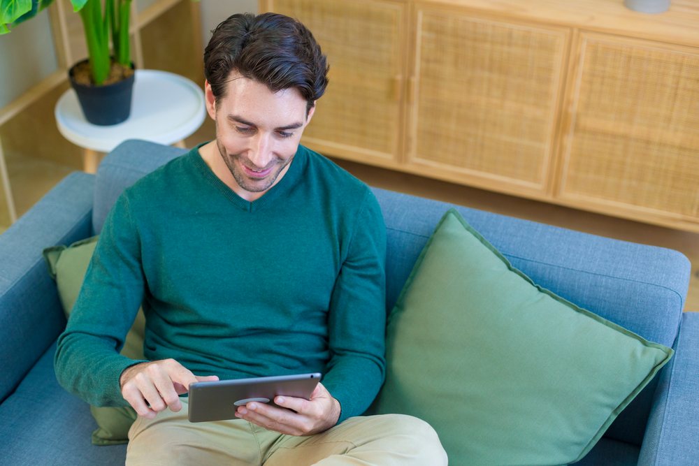 Young man sits comfortably on a couch using a tablet in a modern, well-furnished home interior, technology, young adult.
