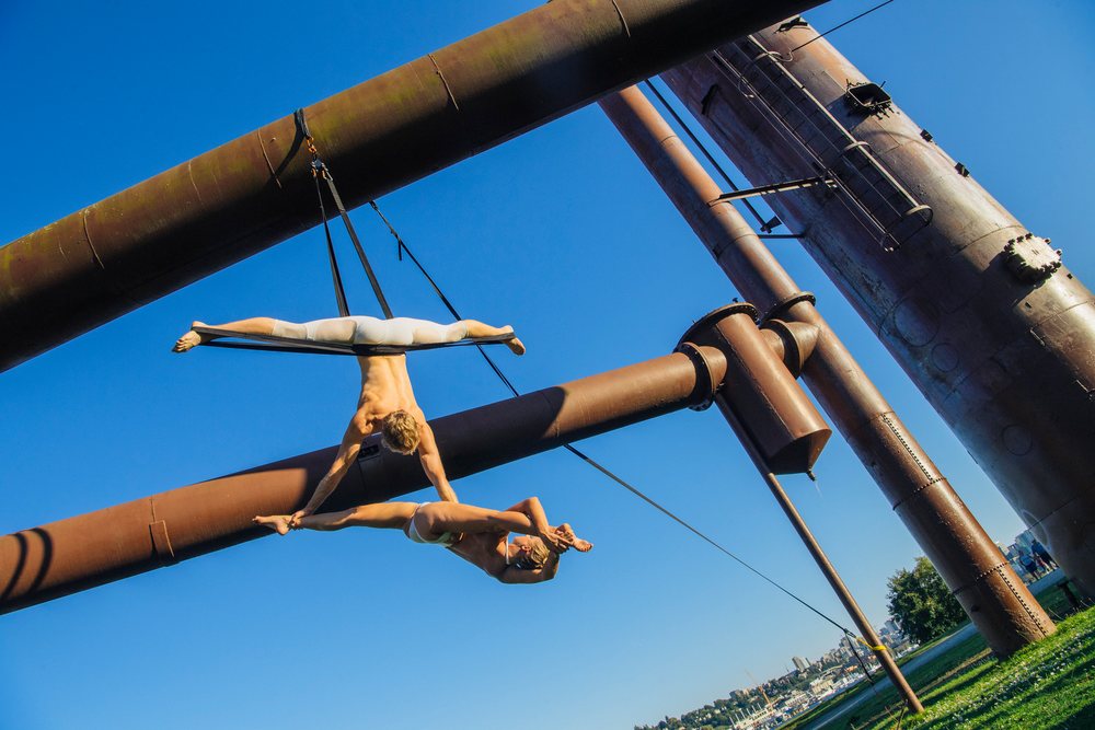 Two aerialists perform acrobatic poses suspended outdoors from a rusted metal structure in a Seattle park, with spectators watching, WA, USA.