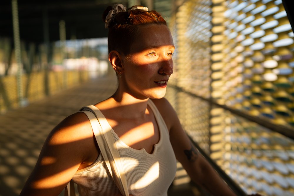 A woman with red hair and a tattoo stands on an outdoor urban bridge bathed in patterned sunlight, Santiago De Compostela, Spain.