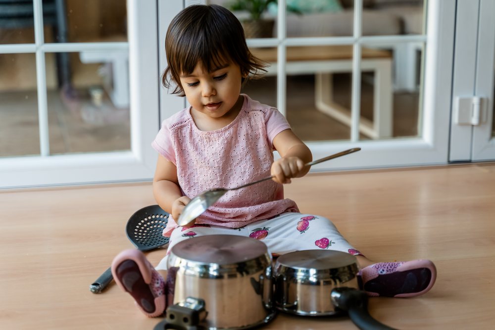 Toddler playing with two metal pots, a plastic strainer, and a spoon on a wooden floor indoors at home, kids, Spain.