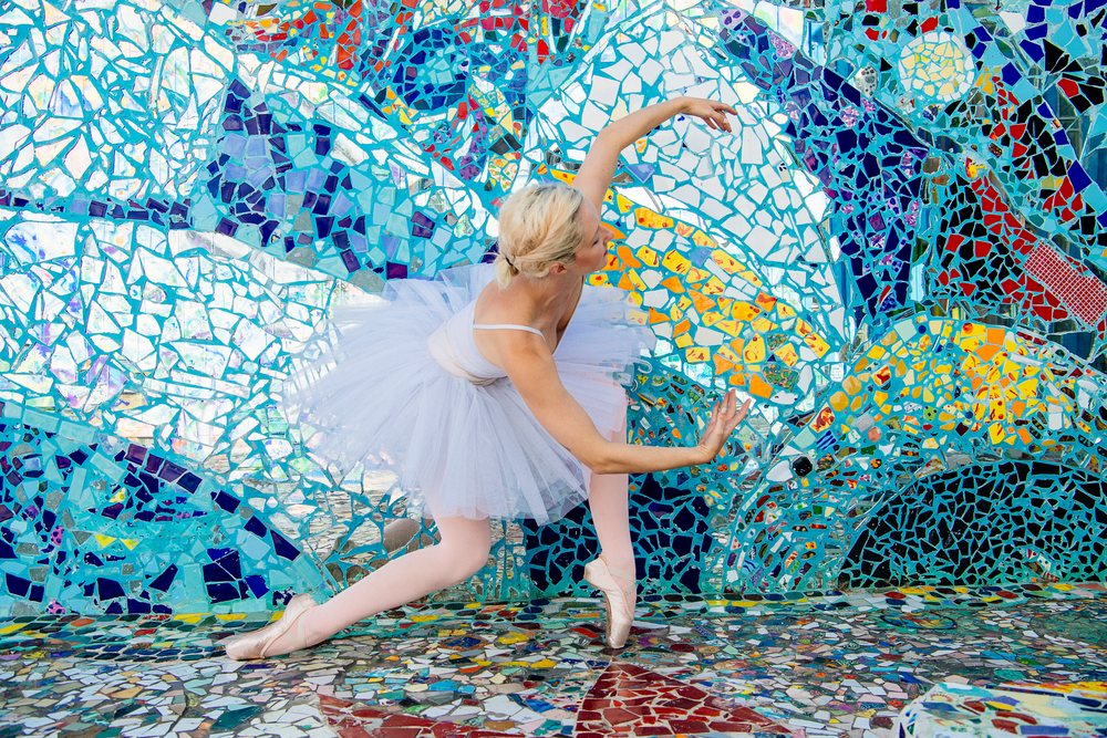 Ballet dancer wearing a tutu and pointe shoes poses in front of a colorful tiled mosaic wall outdoors, CA, USA.