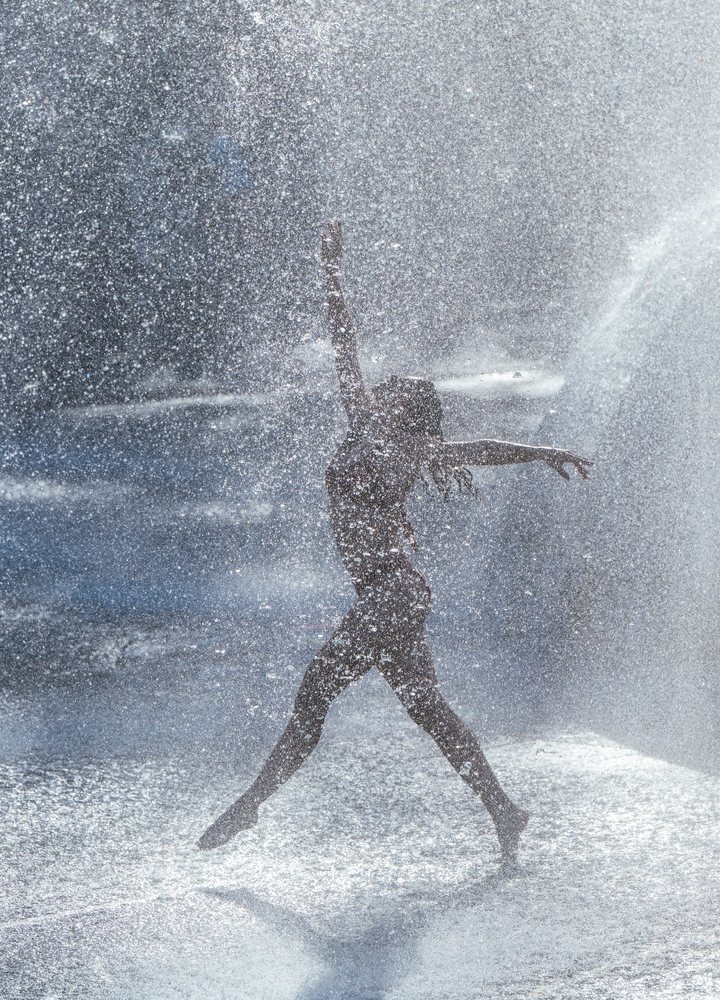 Woman jumping and posing dynamically in water spray outdoors, partner, acrobatics, yoga, USA.