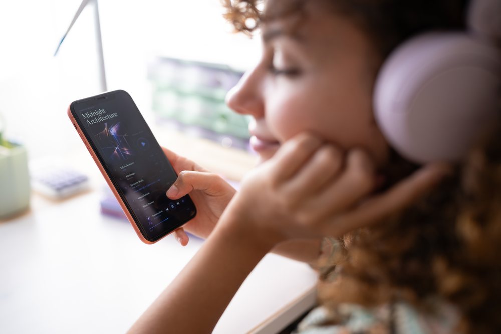 Woman with curly hair wearing headphones and listening to music on a smartphone indoors in a relaxed home setting, Spain.