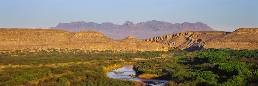 Desert river flows through green vegetation in a valley with distant mountains under a clear blue sky on a sunny day.