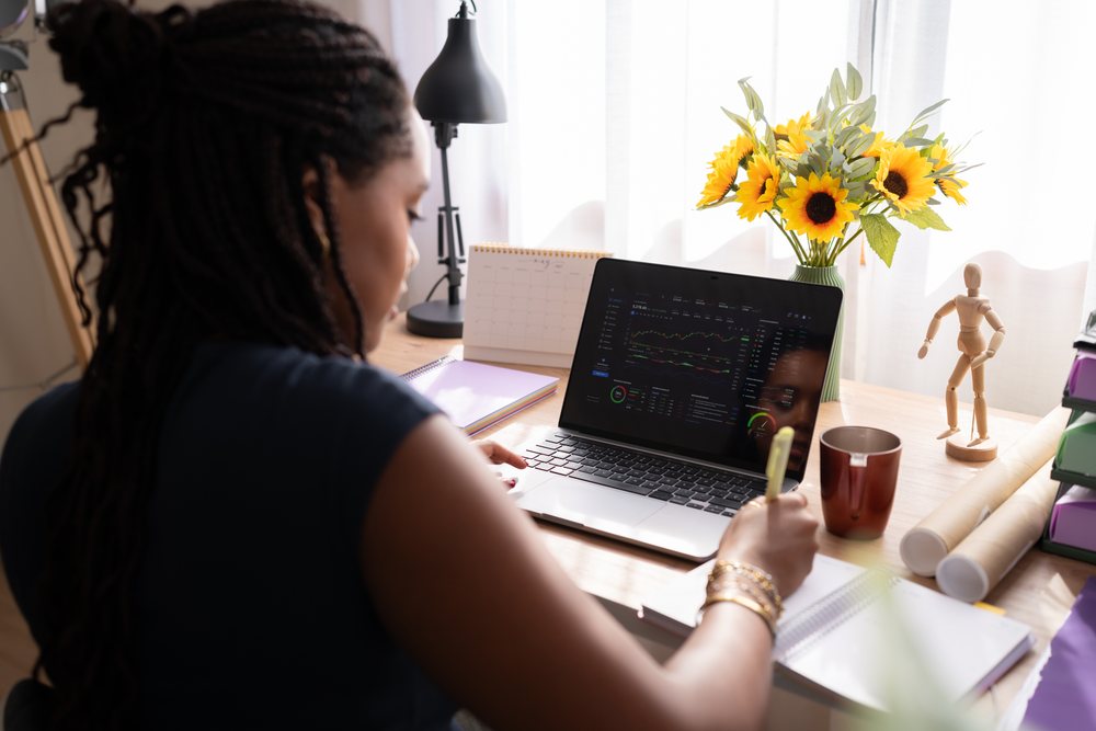 A Black woman works and writes notes at a desk with a laptop displaying financial data in a home office with natural light, sunflowers, and a wooden mannequin, remote work, student, Spain.