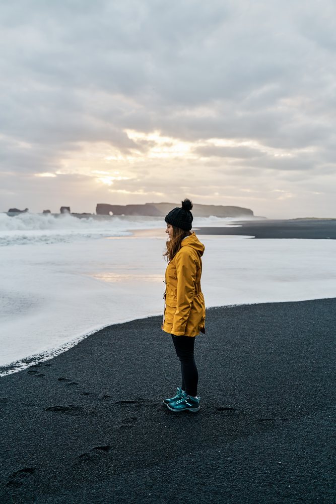 A white woman stands on a black sand beach at sunset in Iceland