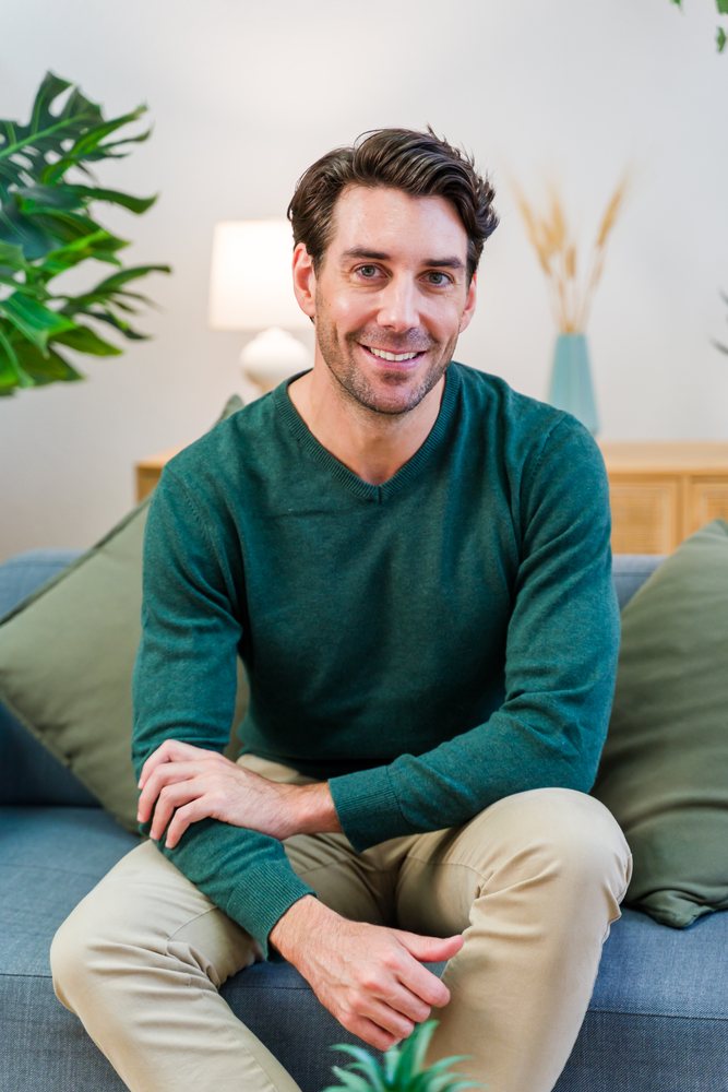 Man wearing green sweater and khaki pants sits smiling on a couch in a modern living room with plants and soft lighting, Spain.
