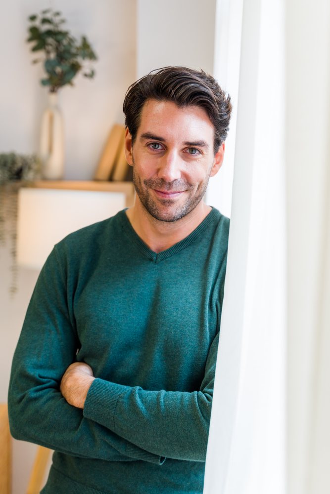 Man in green sweater stands indoors beside a white curtain in a relaxed home setting, Spain.