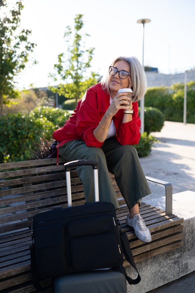 Woman sitting on bench with coffee and luggage outdoors in urban park, Spain.