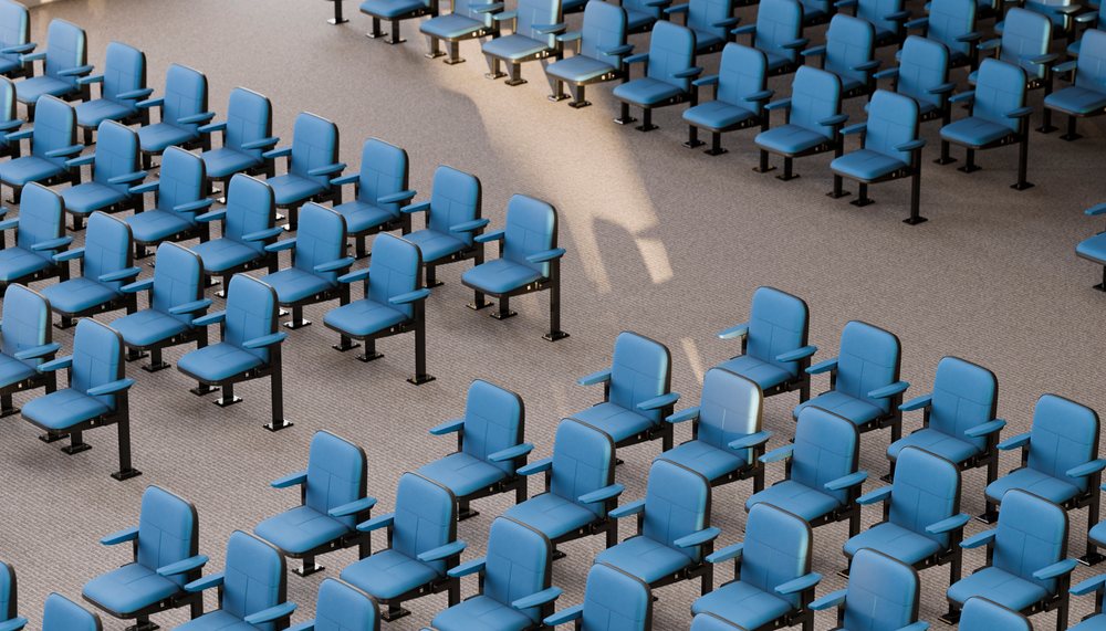 Blue chairs with armrests are arranged in rows on a carpeted indoor floor with natural light casting shadows.
