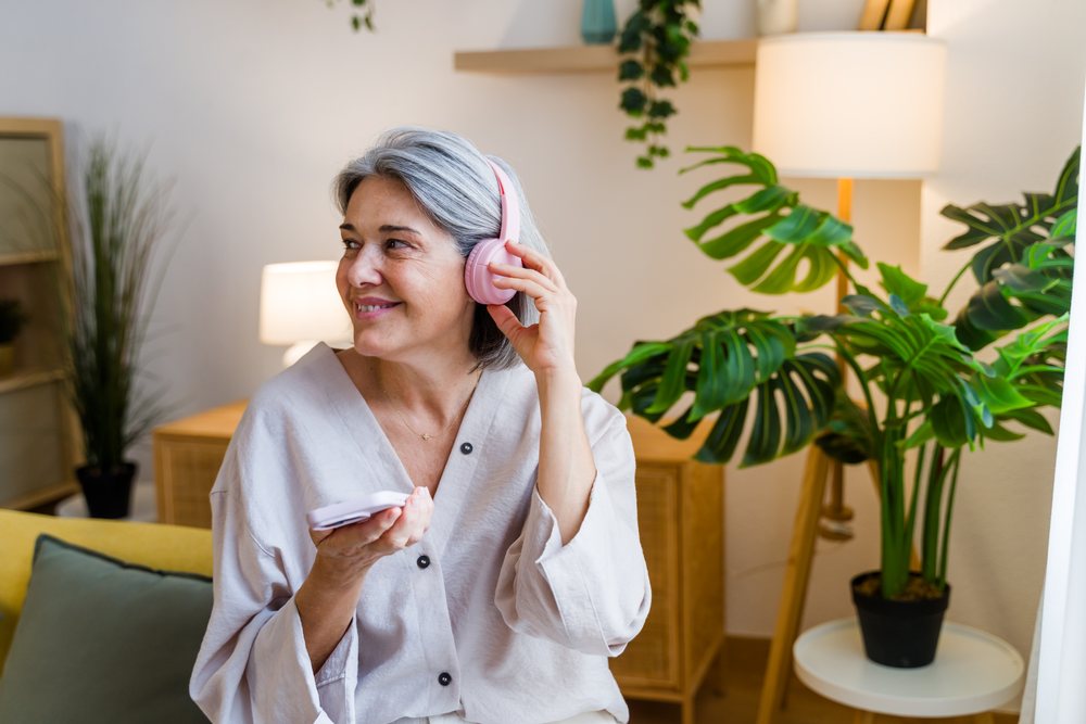 Smiling woman with grey hair wearing pink headphones holds a tablet indoors, Spain.