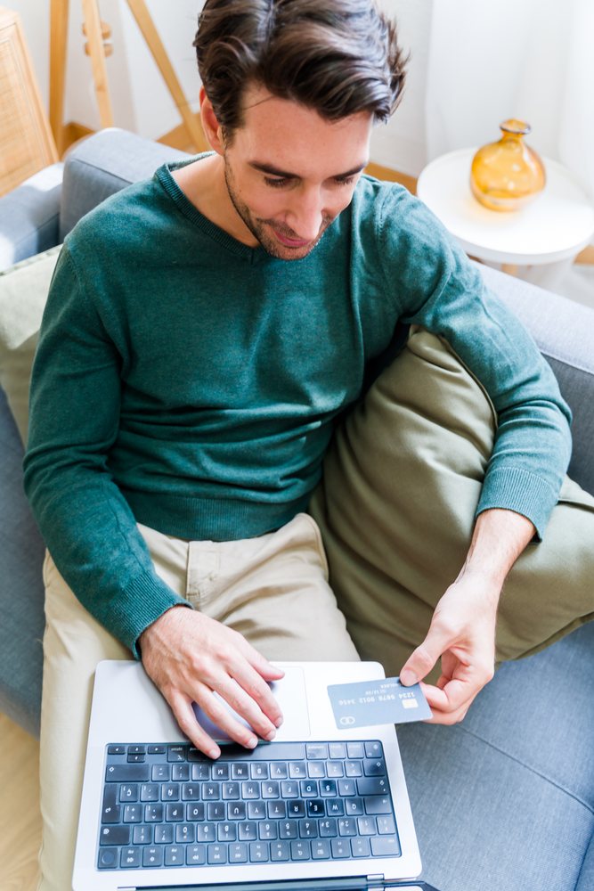 Man wearing a green sweater and beige pants holds a credit card while using a laptop on a grey couch in a living room, Spain.