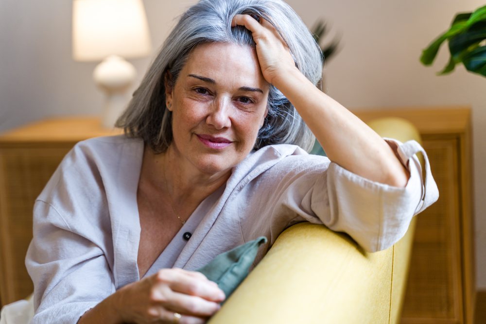 Woman with gray hair smiling and relaxing on a yellow sofa indoors at home, Spain.