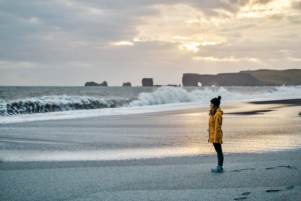 A white woman stands on a black sand beach with waves and sea stacks