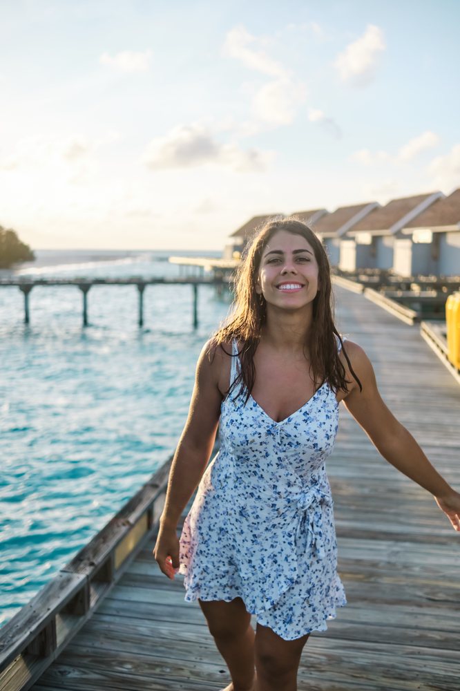 A smiling woman in a white floral summer dress walks along a wooden pier by overwater bungalows in the Maldives at sunset.