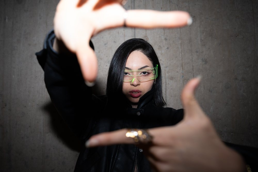 Woman with dark hair and glasses poses indoors framing her face with her hands against a concrete wall, Madrid, Spain.