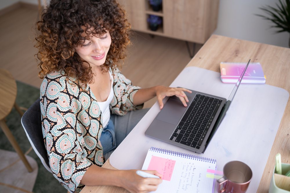 Girl with curly hair works on a laptop and writes notes at a wooden desk in a modern home setting, Spain.