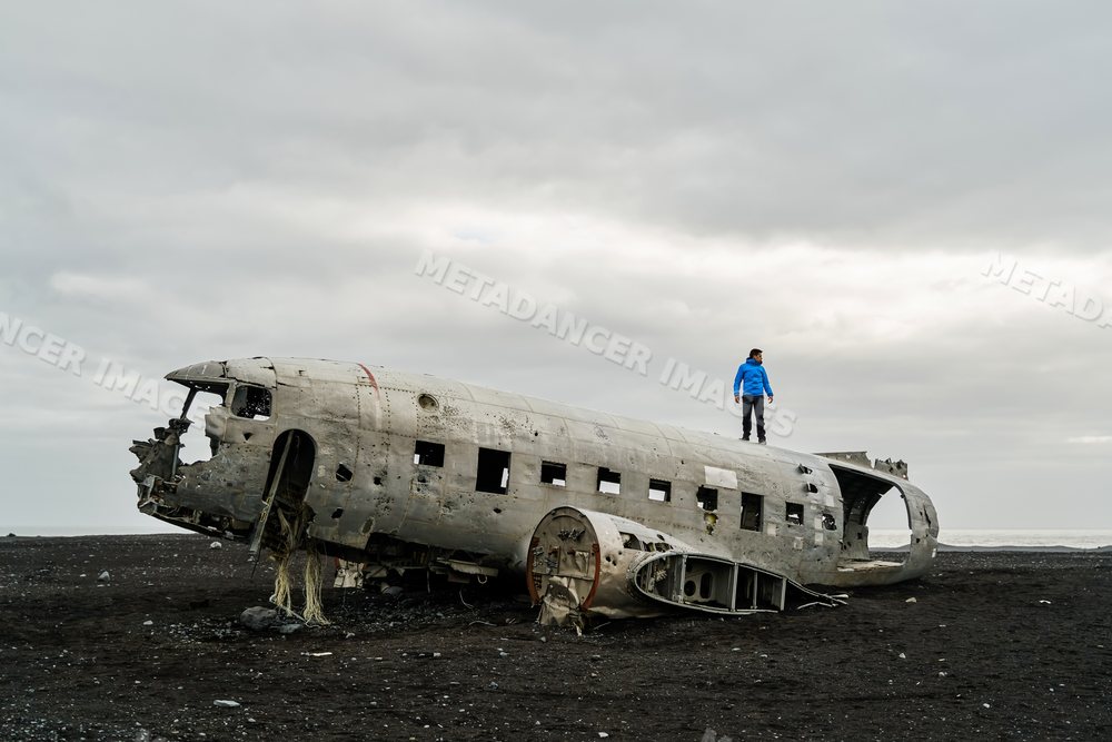 A white man explores the wreck of a crashed plane on a black sand