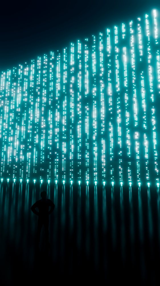 Silhouette of a person facing a large digital installation of vertical glowing blue lights against a dark background with floor reflections.