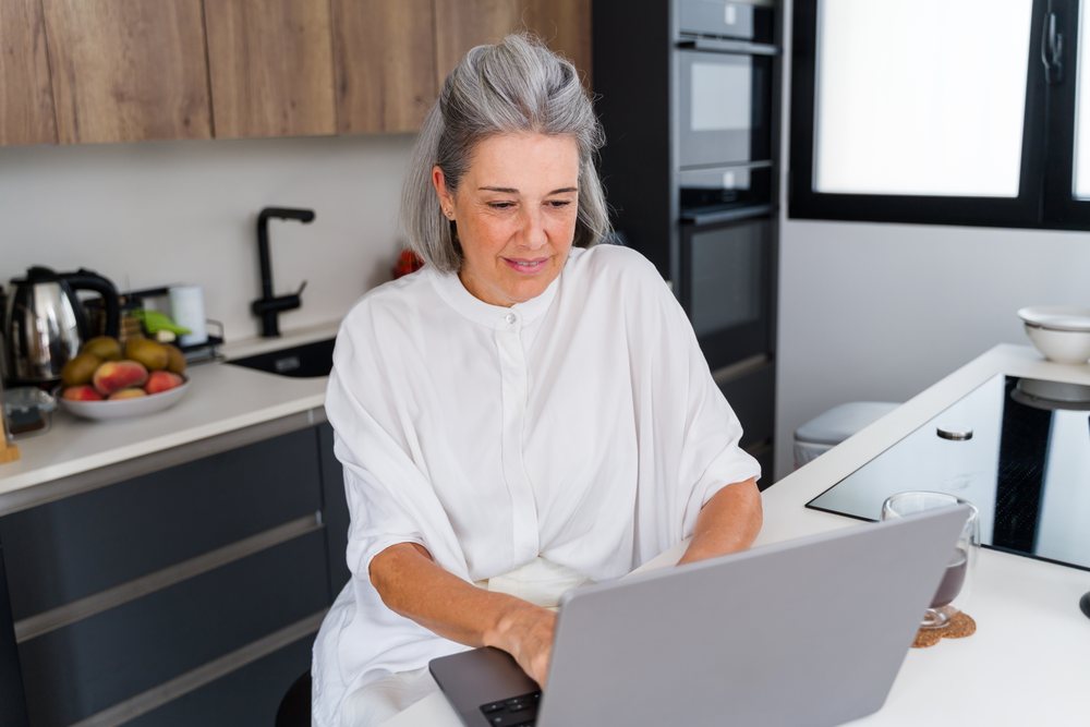 Woman with gray hair working on a laptop at a kitchen counter in a modern kitchen, Spain.