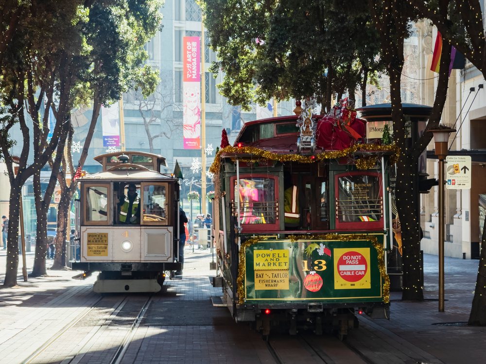 Two decorated San Francisco cable cars travel along Market Street under trees wrapped with holiday lights in downtown, landscape, cityscape, city life, California, USA.