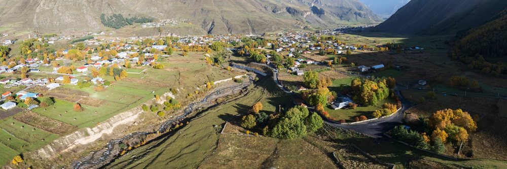 Aerial view of Sioni Village in Georgia's Kazbegi mountainous valley showing buildings, autumn trees, river, and surrounding mountains, Kazbegi Region.