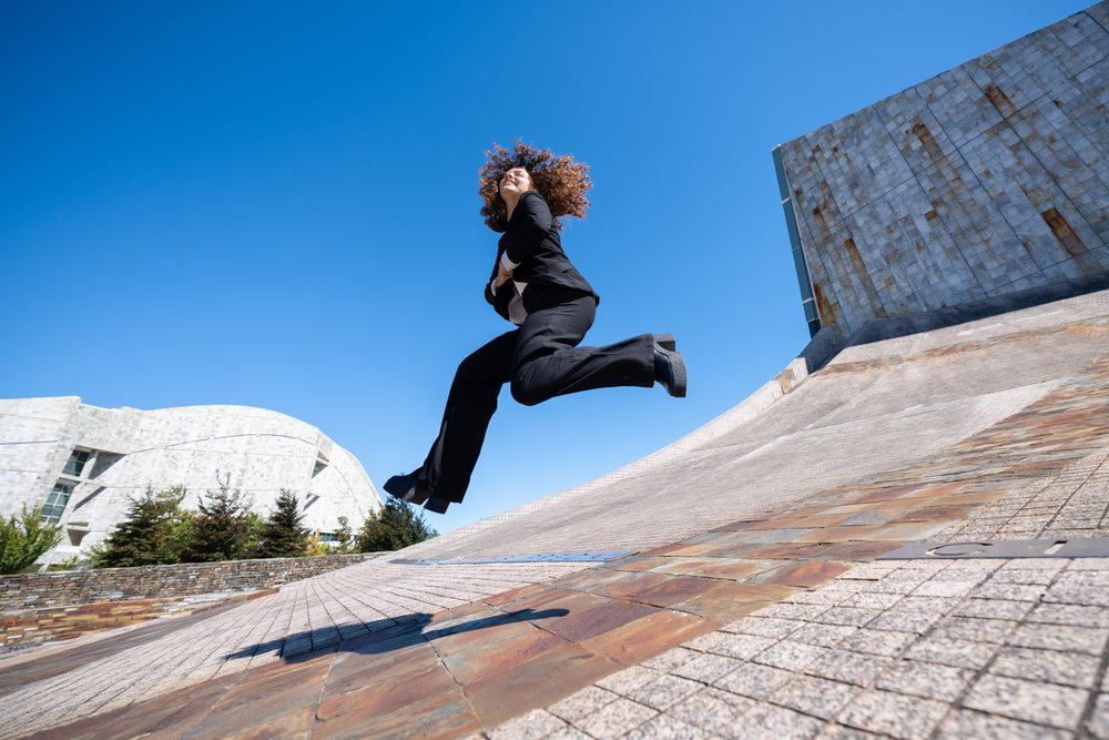 Woman jumping energetically near a modern curved concrete wall with blue sky in Santiago de Compostela, businesswoman, traveling executive, Spain, Antiago De Compostela.