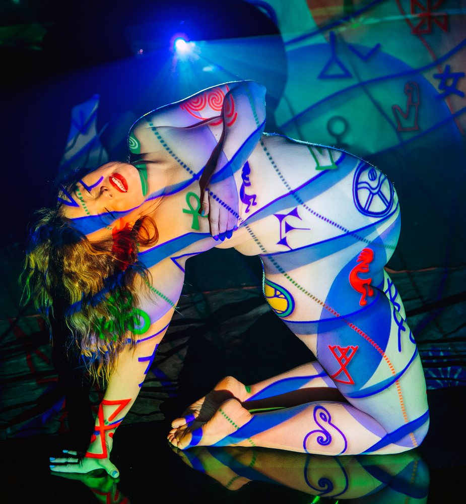 Nude woman kneels in a dark studio as colorful symbolic patterns are projected onto her body and the background, pregnancy.
